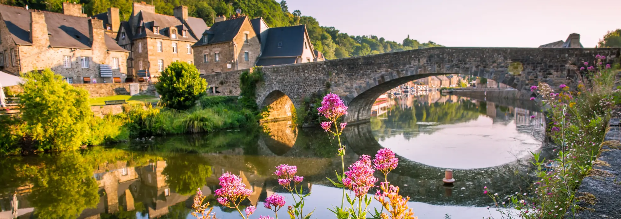 Malerische Steinbrücke in Dinan in der Bretagne, gesäumt von historischen Fachwerkhäusern am Ufer des Flusses Rance mit bunten Blumen im Vordergrund.