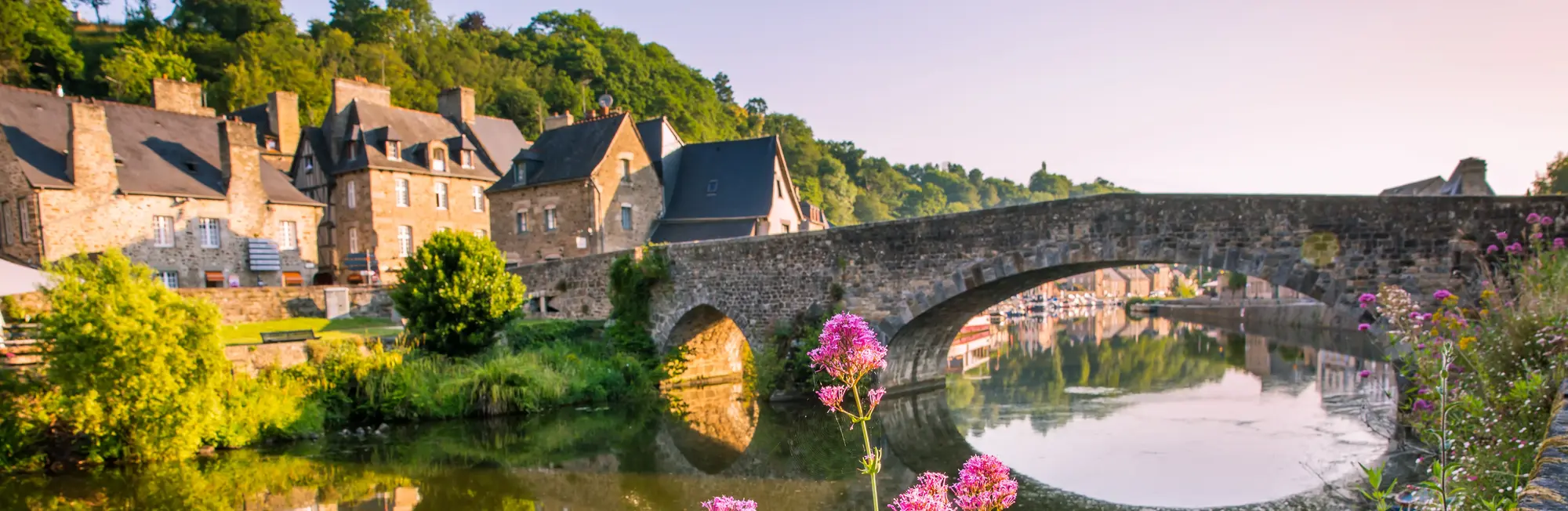 Malerische Steinbrücke in Dinan in der Bretagne, gesäumt von historischen Fachwerkhäusern am Ufer des Flusses Rance mit bunten Blumen im Vordergrund.