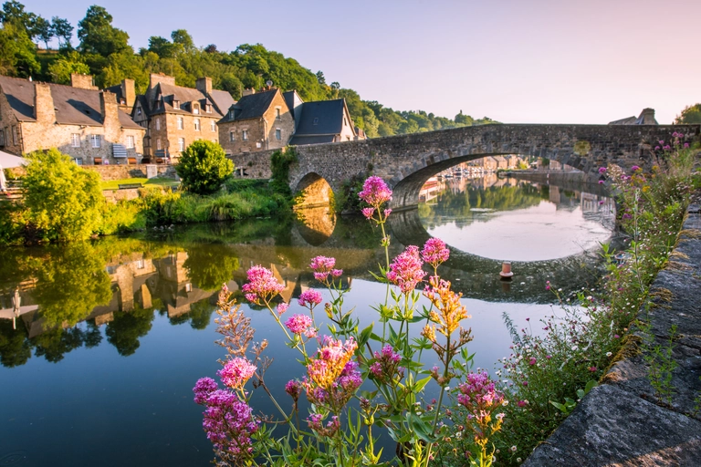 Blick auf das mittelalterliche Städtchen Dinan in der Bretagne, Frankreich. Eine historische Steinbrücke spannt sich über den ruhigen Fluss, der die Häuser am Ufer spiegelnd widerspiegelt. Im Vordergrund blühen pinke Blumen, während warmes Abendlicht die Szene in sanfte Farben taucht.
