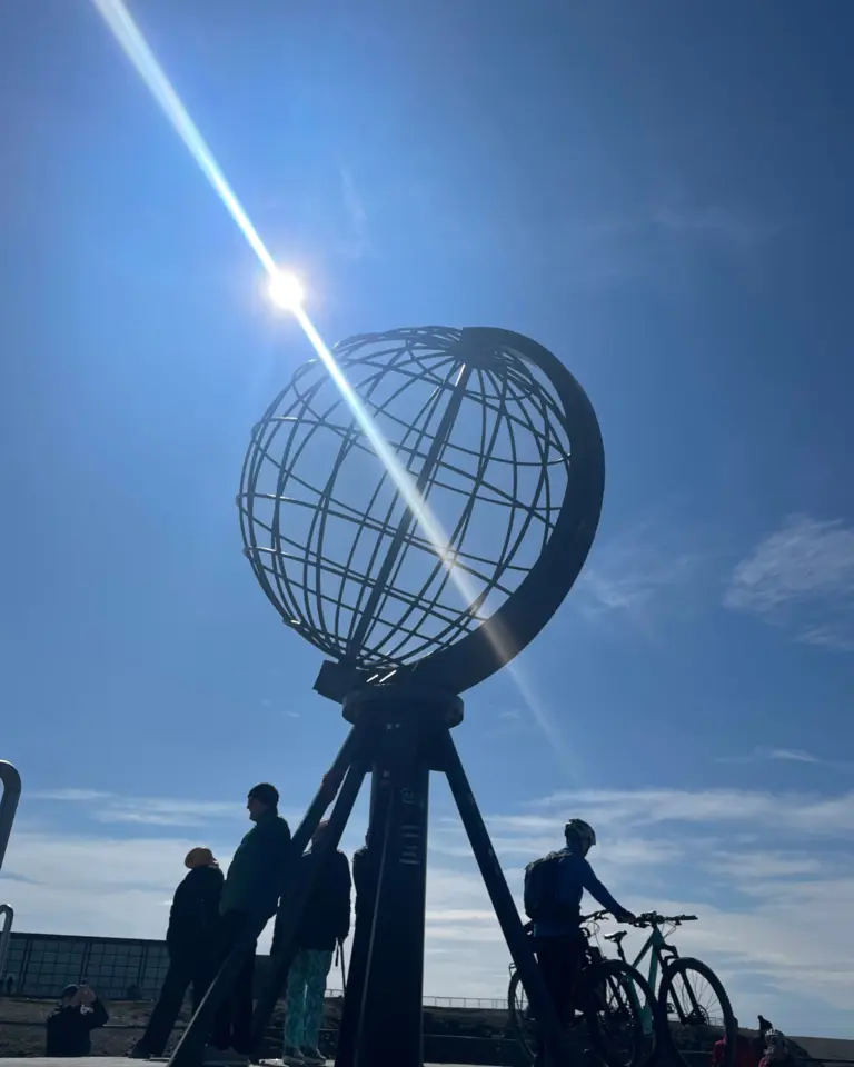 Nordkap Auf diesem Bild ist das berühmte Globus-Denkmal am Nordkap in Norwegen zu sehen, das sich als Silhouette gegen die strahlende Sonne und den blauen Himmel abzeichnet. Man erkennt mehrere Touristen, darunter einen Radfahrer, die sich um das Wahrzeichen am nördlichsten Punkt Europas versammelt haben.
