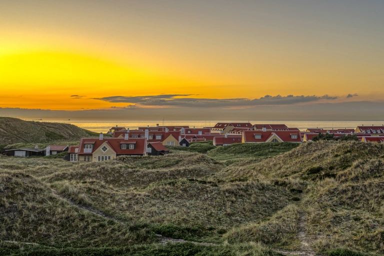 Blick auf die roten Dächer der Häuser von Skagen, eingebettet in sanfte Dünenlandschaft, während im Hintergrund ein goldener Sonnenuntergang über dem Meer leuchtet.