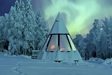 Nordlichter über verschneiter Kota-Hütte in Lappland Eine verschneite, pyramidenförmige Kota-Hütte steht in einer Winterlandschaft in Lappland, umgeben von vereisten Bäumen. Am klaren Nachthimmel leuchten grün schimmernde Nordlichter über dem Dach.
