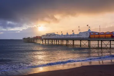 Der Brighton Palace Pier an der Südküste Englands ragt ins Meer hinaus. Die beleuchtete Seebrücke mit Fahrgeschäften und Gebäuden spiegelt sich im Wasser, während die Sonne bei dramatischem Abendhimmel über dem Strand untergeht.