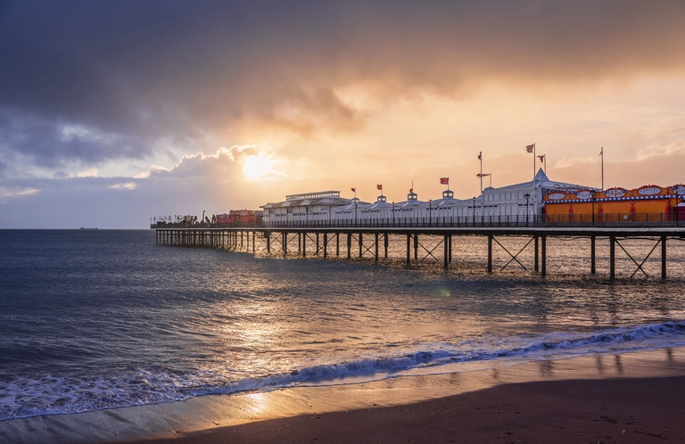 Der Brighton Palace Pier an der Südküste Englands ragt ins Meer hinaus, während die Sonne untergeht. Das warme Licht spiegelt sich im Wasser, und die beleuchtete Seebrücke zeigt die typische Küstenatmosphäre eines beliebten Reiseziels in Europa.